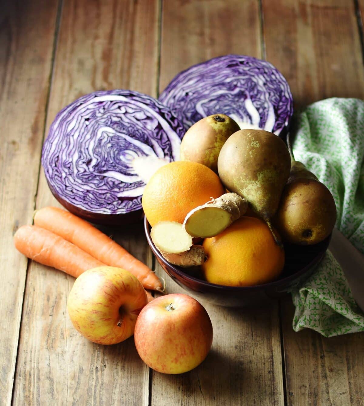Raw apples, carrots and halved red cabbage on top of wooden surface with pears, oranges and ginger inside purple bowl and green cloth to the right.