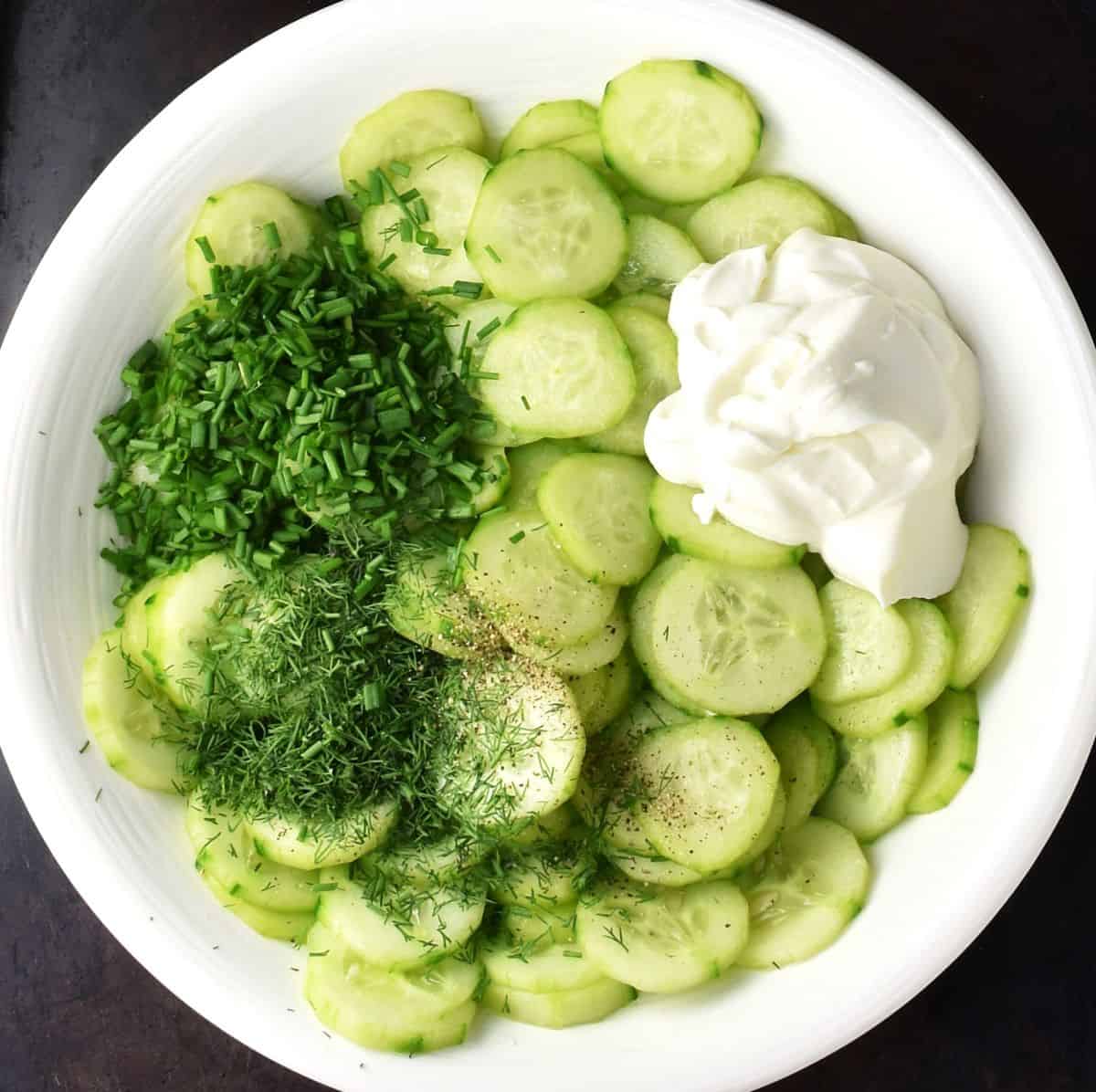 Cucumber slices with fresh dill and chives and sour cream in white bowl.
