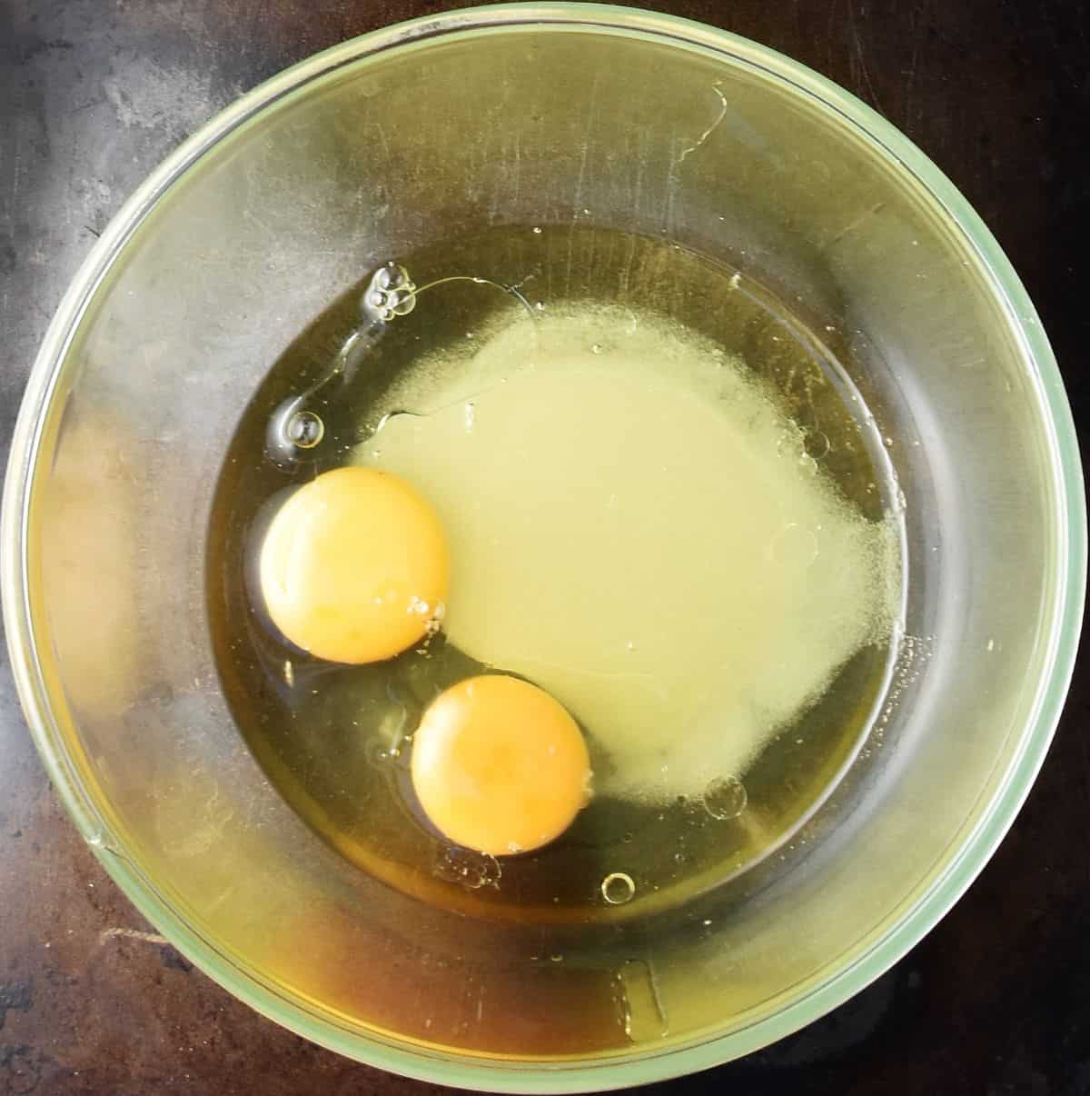 Top down view of 2 eggs with sugar and oil in glass bowl.
