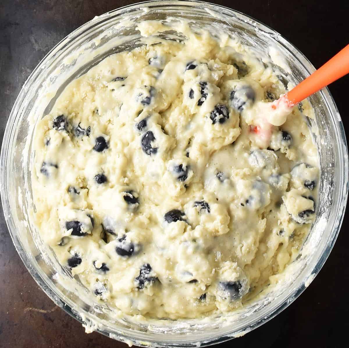 Top down view of blueberry muffin cake batter in glass bowl with orange spatula.