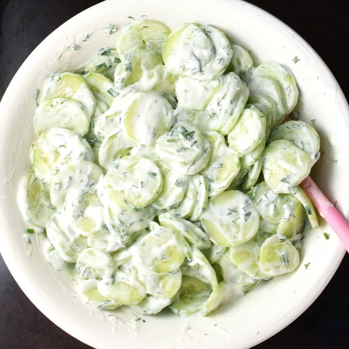 Making Polish cucumber salad with sour cream in white bowl with pink spatula.