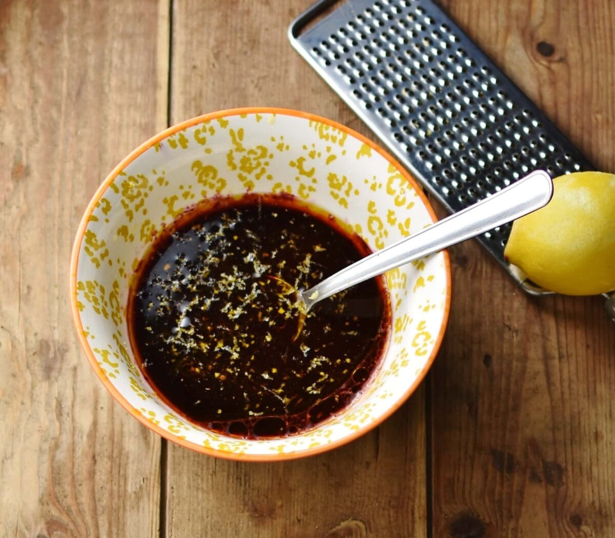 Blueberry vinaigrette ingredients with spoon in yellow-and-white patterned bowl, with lemon and zester in top right corner.