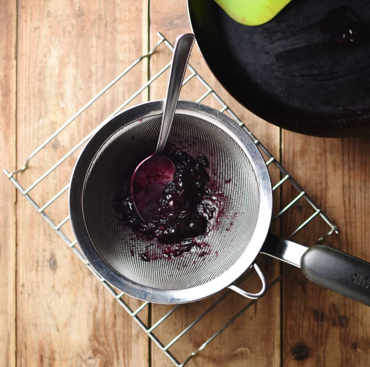 Blueberry pulp with spoon inside sieve on top of saucepan on cooling rack, with pan in top right corner,