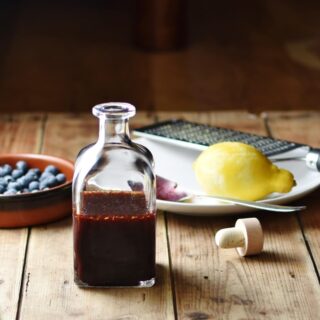 Blueberry vinaigrette in bottle, with blueberries in brown dish, lemon, zester and spoon on top of white plate and bottle top in background.