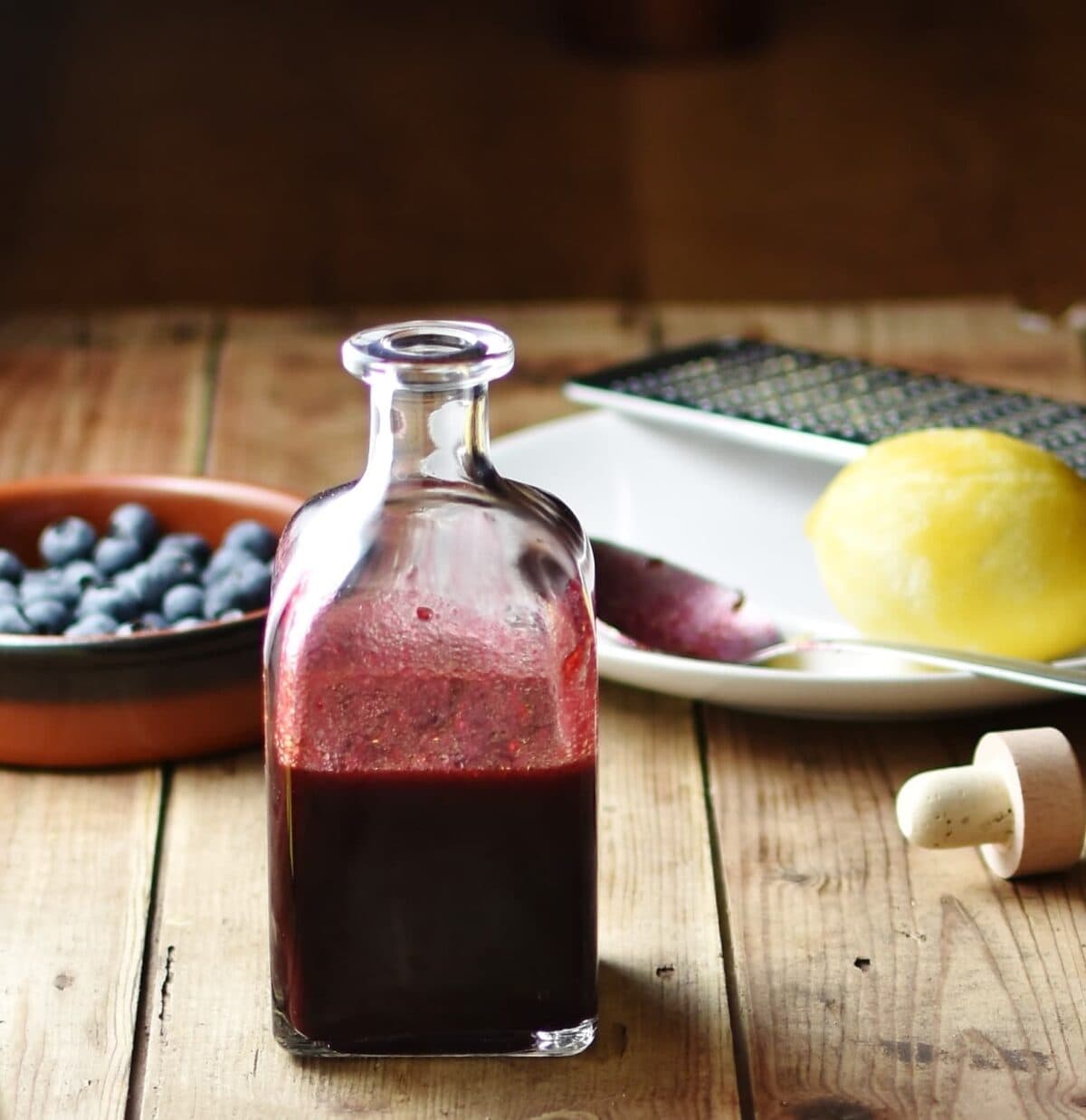 Blueberry dressing in bottle, with blueberries in brown dish, lemon, zester and spoon on top of white plate and bottle top in background.