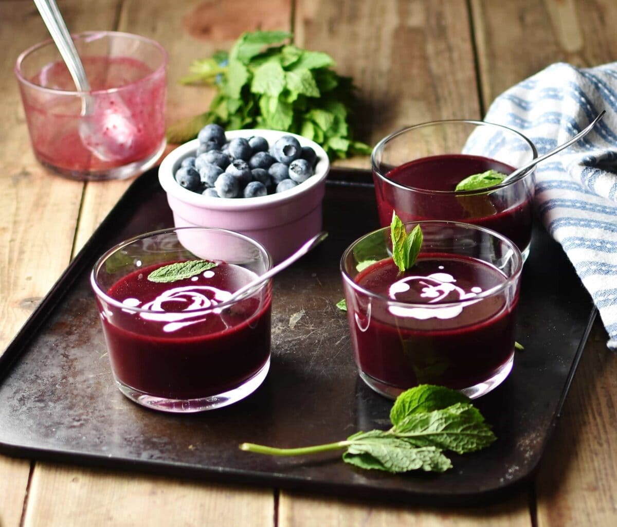 Side view of fruit soup in 3 glasses with fresh mint and blueberries in pink cup on top of tray,with mint, blue cloth and dish with spoon in background.