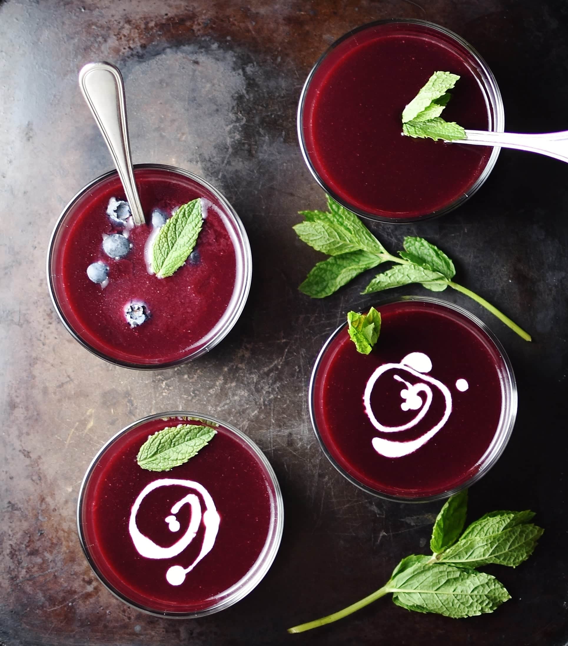Top down view of fruit soup in 4 glasses with 2 spoons, mint leaves and swirls of cream on top of dark surface.