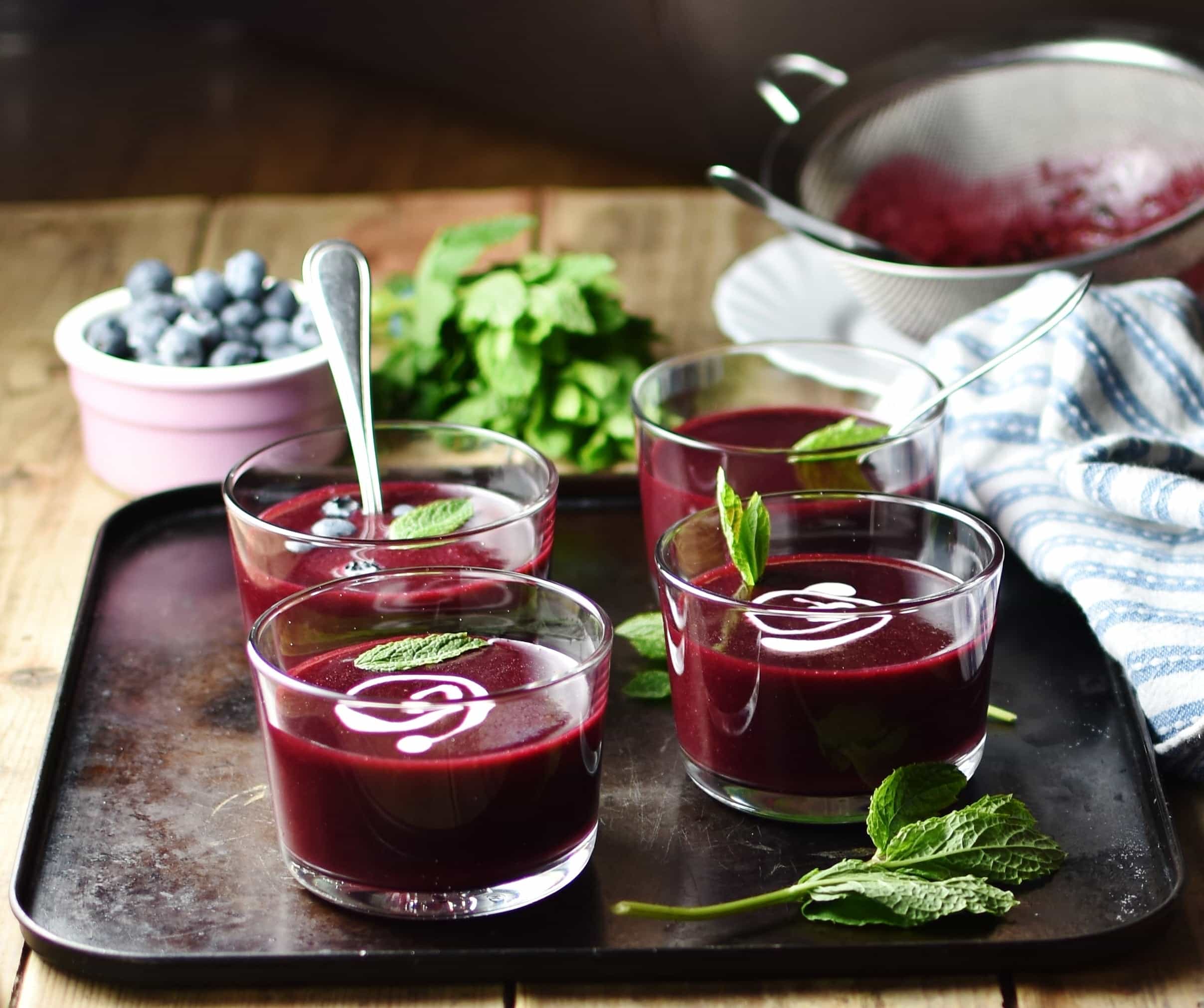 Side view of fruit soup in 4 glasses with spoon, mint leaves and swirls of cream, blue cloth, sieve, mint and blueberries in pink dish in background.