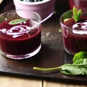 Side view of fruit soup in 2 glasses with spoon, mint leaves and swirls of cream, blueberries in pink dish and fresh mint in background.