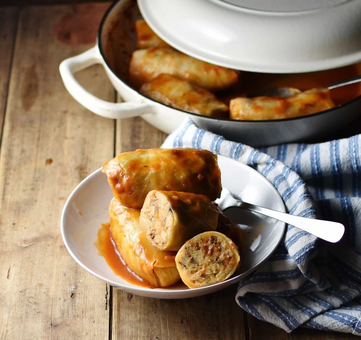 Side view of cabbage rolls inside white bowl with fork, wrapped in blue stripy cloth, with cabbage rolls inside white dish with lid in background.