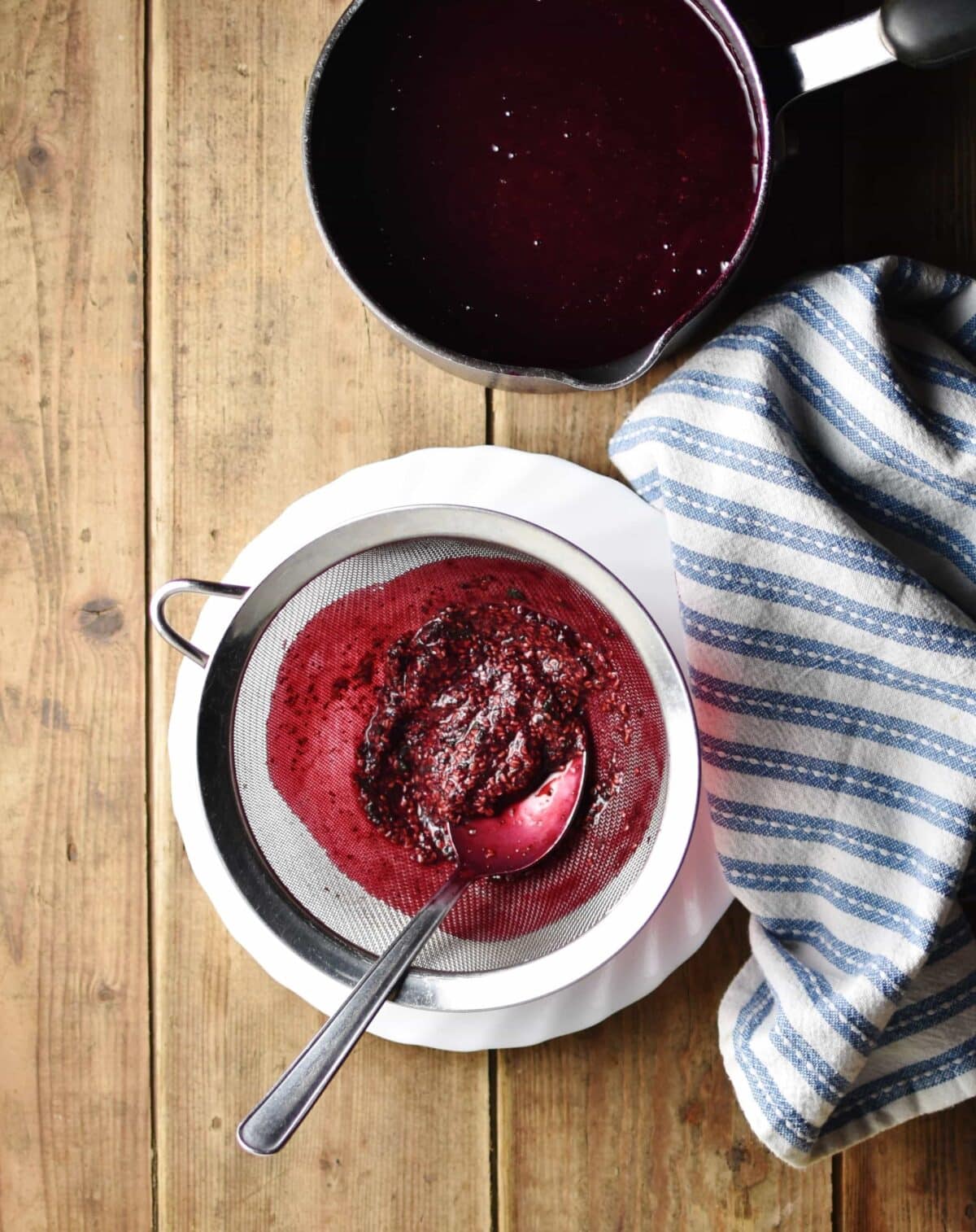 Top down view of fruit seeds inside fine sieve with spoon on top of white plate with stripy blue-and-white cloth and fruit soup in saucepan in background.