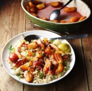 Peaches and quinoa with lemon wedges, nuts, herbs and large spoon on top of white plate, with green oval dish with cooked peaches and juices in background.