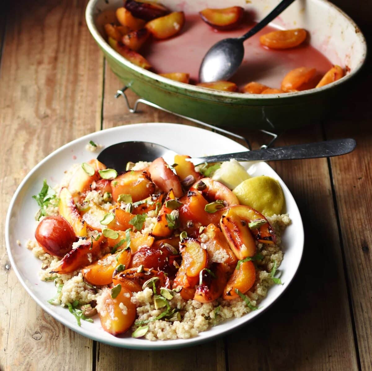 Peaches and quinoa with lemon wedges, nuts, herbs and large spoon on top of white plate, with green oval dish with cooked peaches and juices in background.
