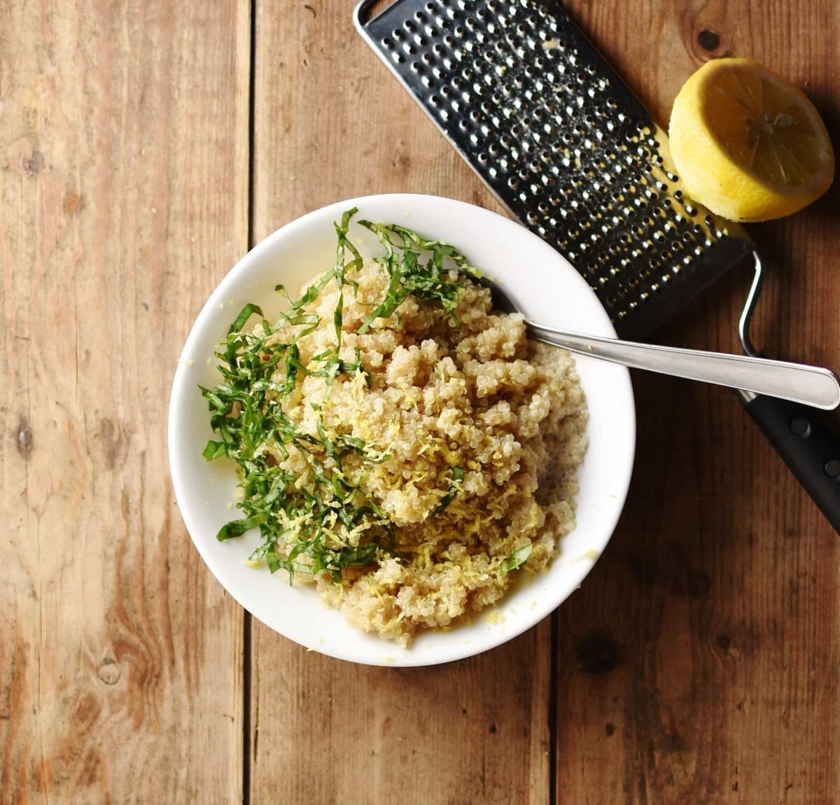 Cooked quinoa with chopped herbs and spoon inside white bowl, with lemon half on top of zester in background.