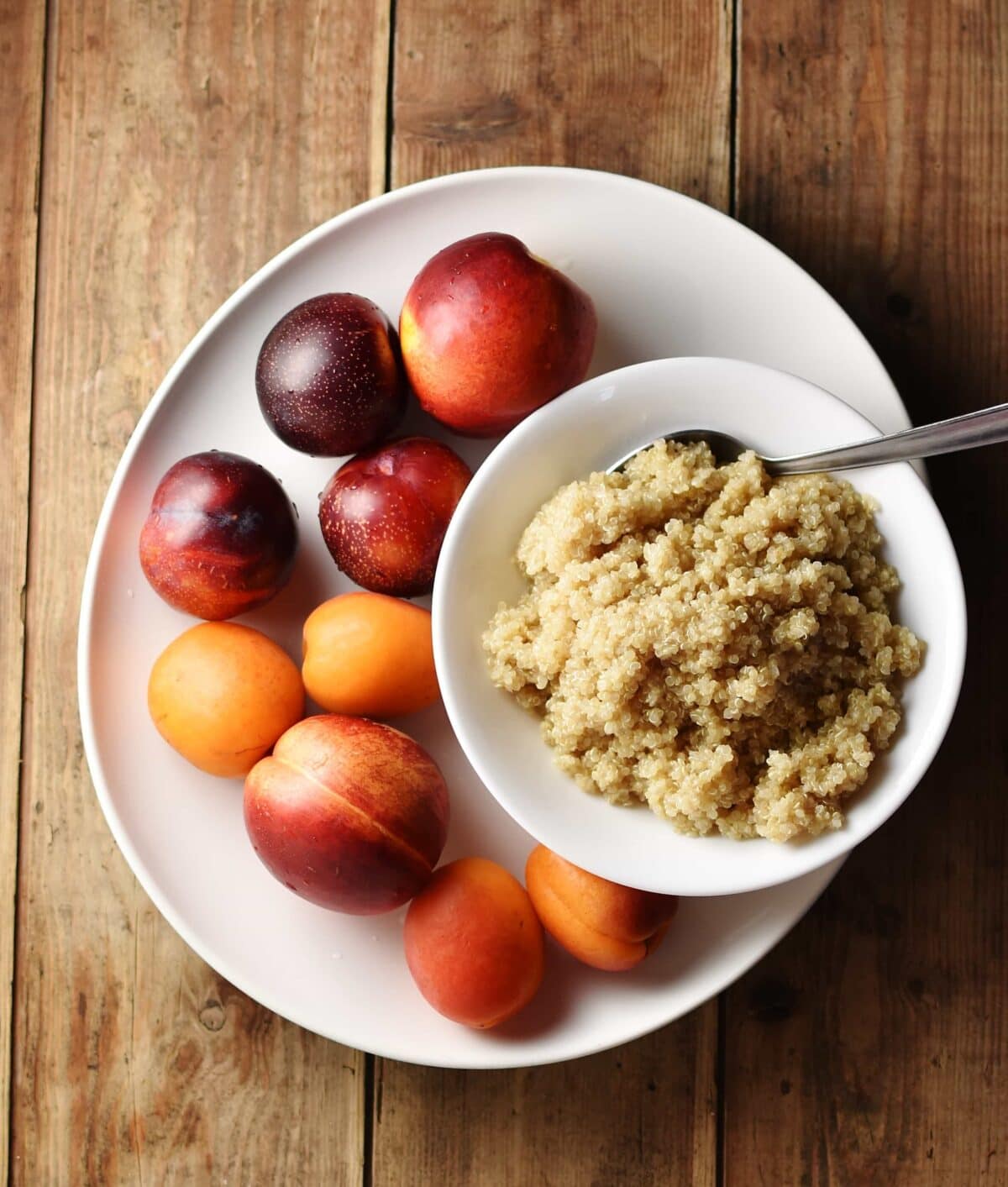 Nectarines, plums and apricots and cooked quinoa inside white bowl with spoon on top of large white plate.