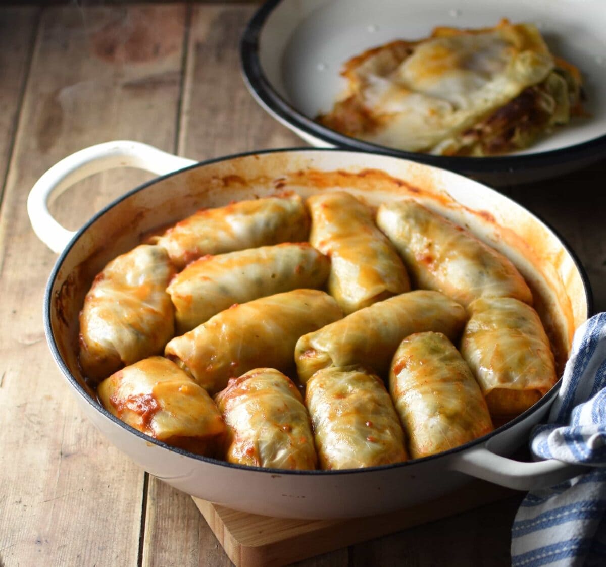 Side view of cabbage rolls in white dish with lid and cabbage leaves on top in background.