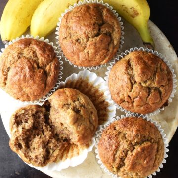 Top down view of banana buttermilk muffins in white paper cases and overripe bananas in background.