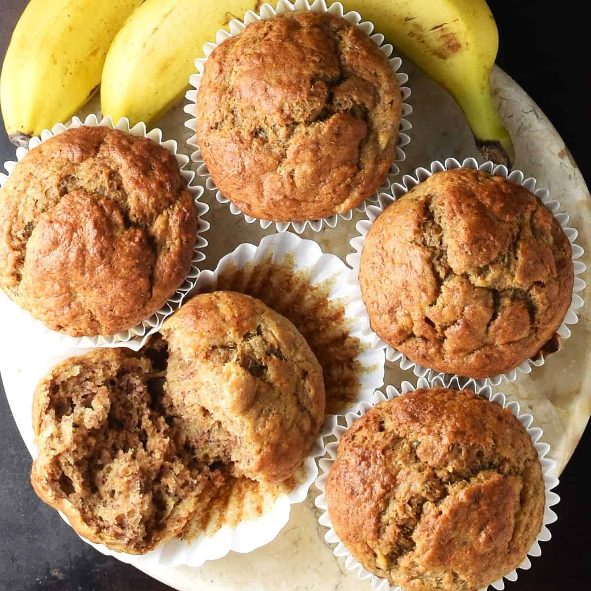 Top down view of banana buttermilk muffins in white paper cases and overripe bananas in background.