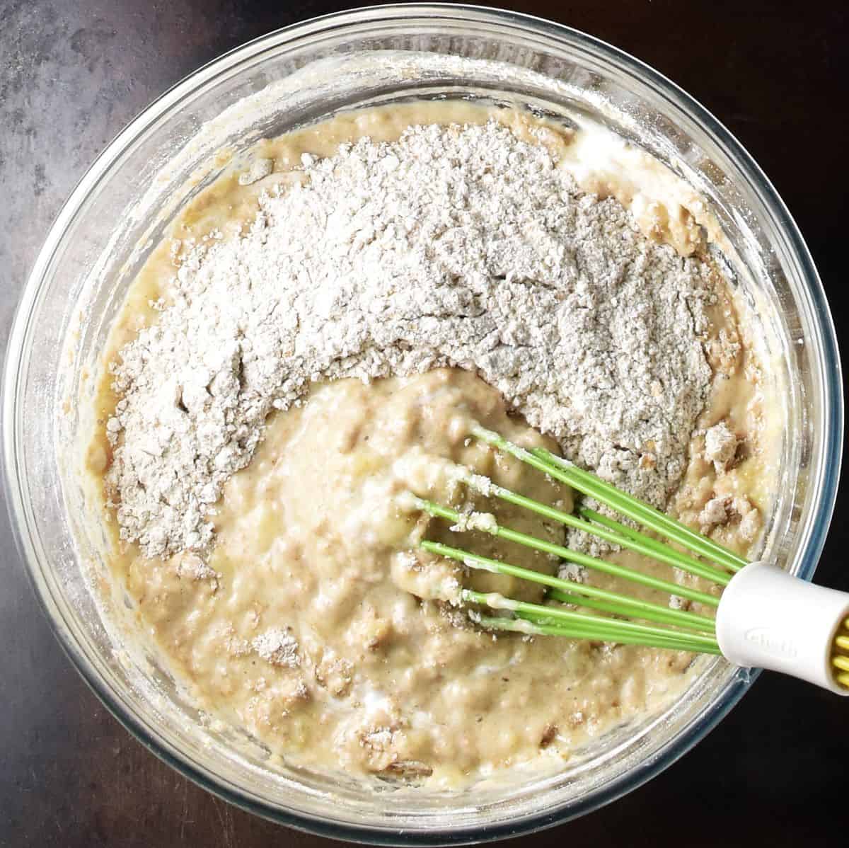 Banana buttermilk muffin batter with flour visible in glass bowl with whisk.
