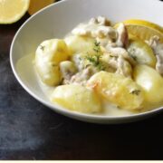 Potato and chicken in creamy sauce in white bowl with lemon and yellow dish in background.
