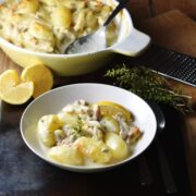 Side view of potato and chicken in creamy sauce in white bowl with yellow casserole dish and halved lemon in background.