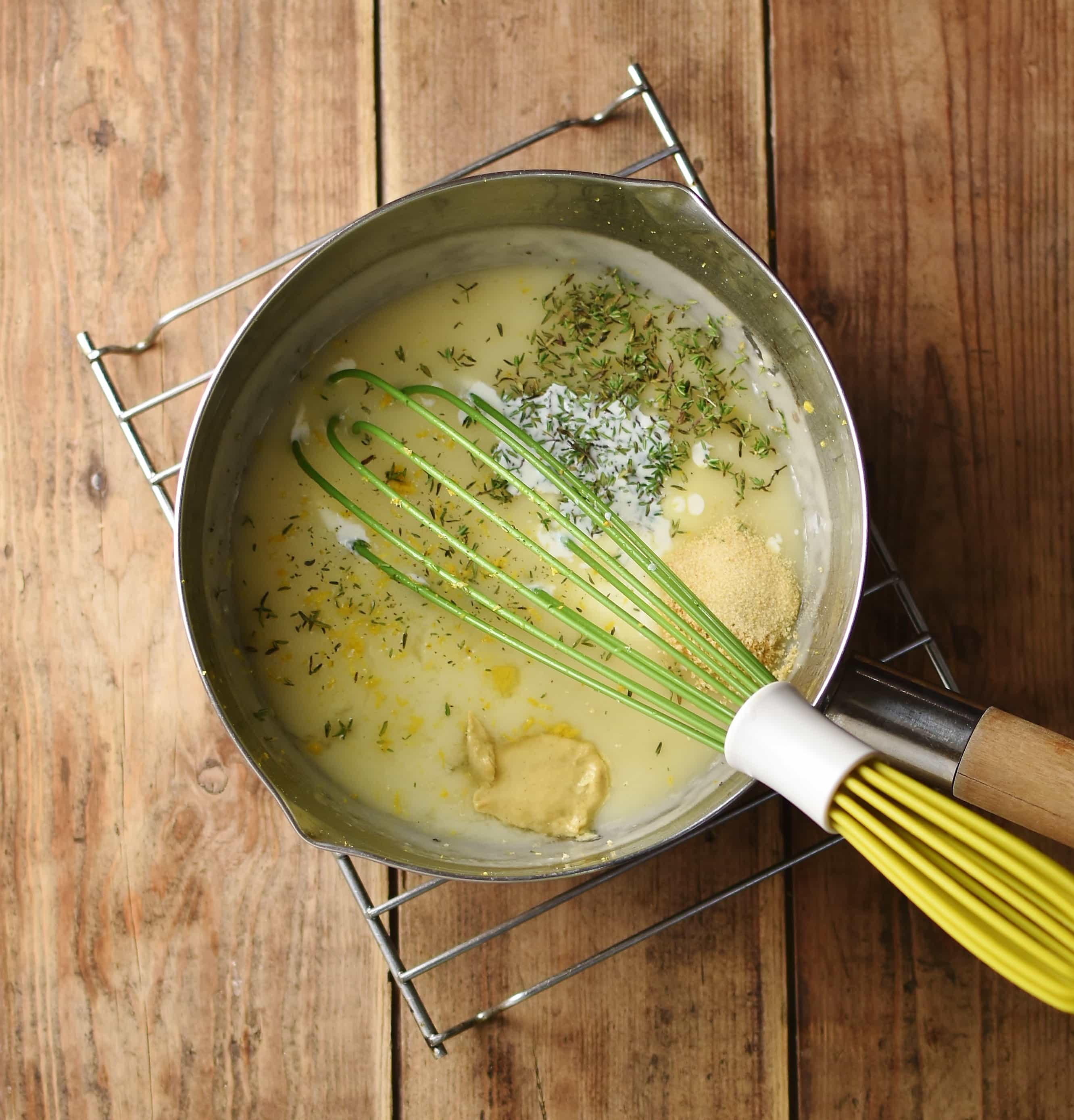 White sauce with mustard, herbs and spices in saucepan with green whisk on top of cooling rack.