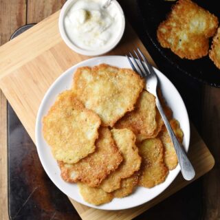 Pancakes with fork on white plate, with yogurt in small dish on top of wooden board, and with pancakes in skillet in top right corner.
