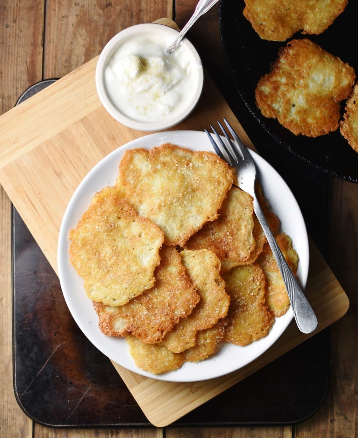Pancakes with fork on white plate, with yogurt in small dish on top of wooden board, and with pancakes in skillet in top right corner.