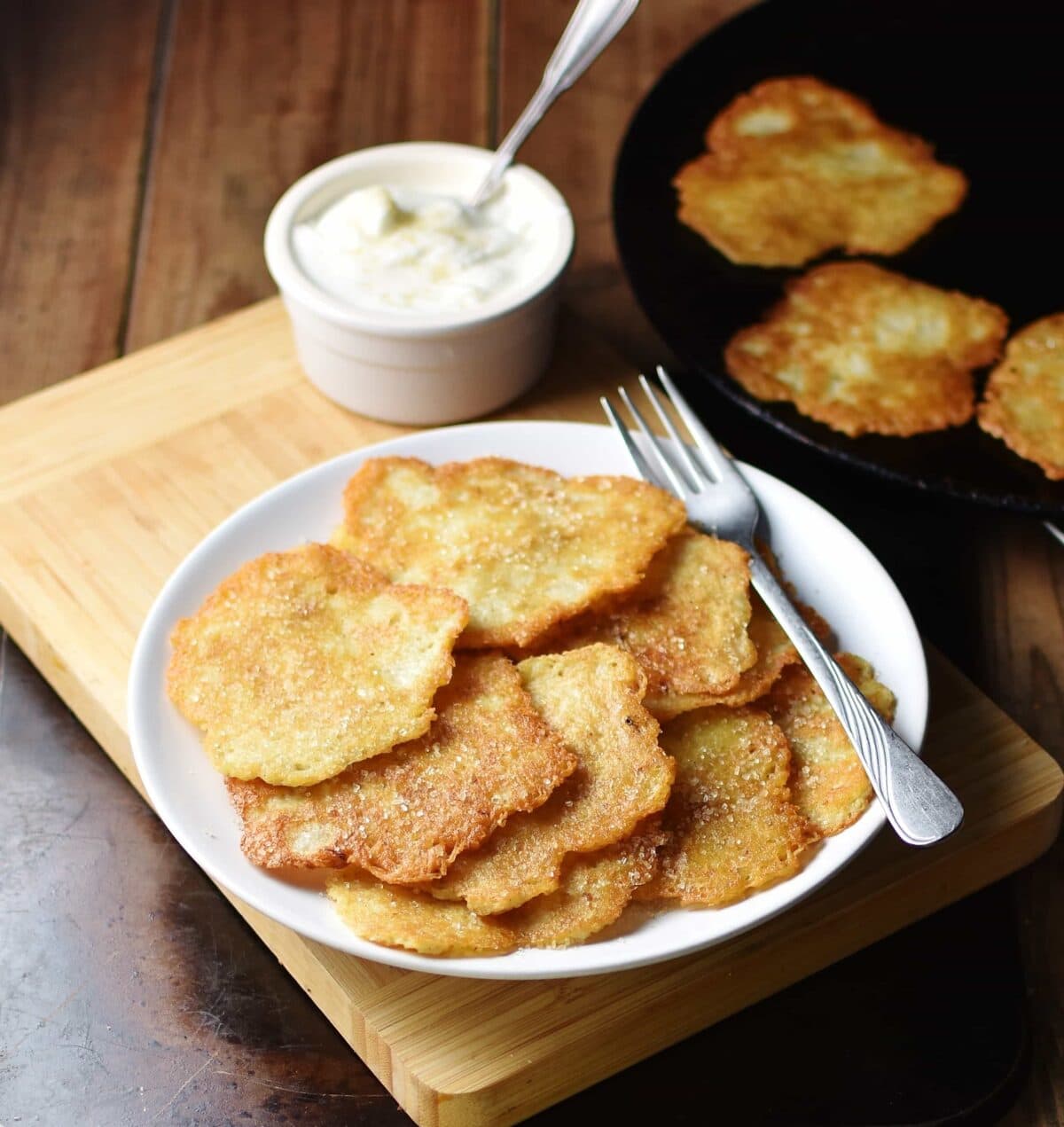 Potato pancakes with fork on top of white plate with yogurt in white dish with spoon and more pancakes in skillet in background.