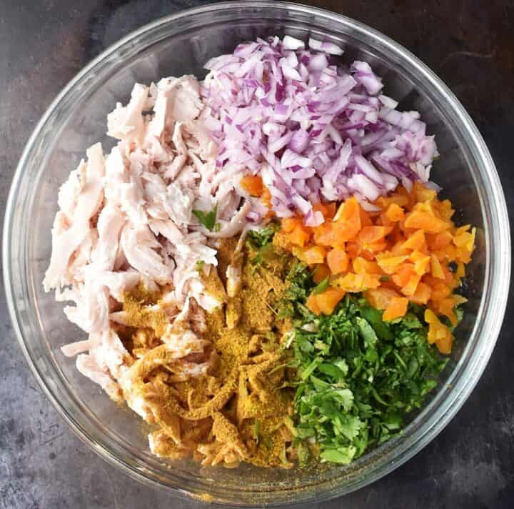 Top down view of shredded chicken, onion, herbs and apricots in glass bowl.