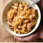 Bigos with spoon in green bowl held by 2 hands, with more stew in pot in top right corner.