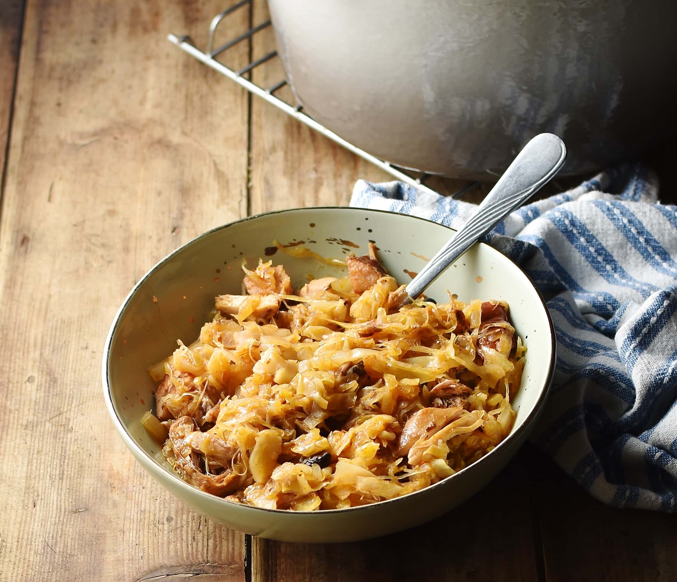 Sauerkraut bigos in green bowl with spoon, stripy blue-and-white cloth and grey pot in background.