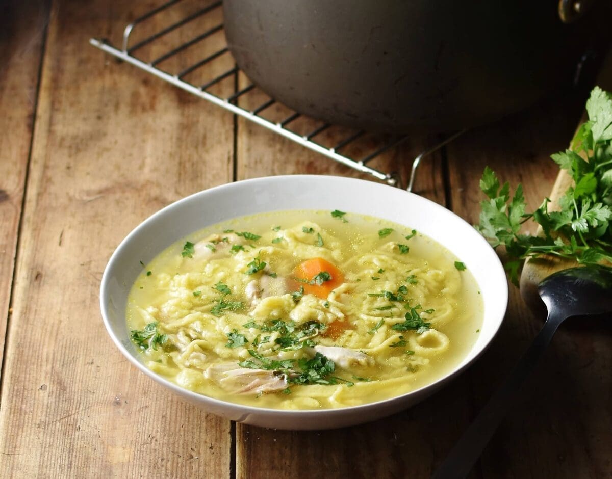 Side view of chicken egg drop noodle soup in white bowl with pot on cooling rack in background.