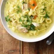 Egg drop noodles chicken soup with carrot and chopped herbs in white bowl.