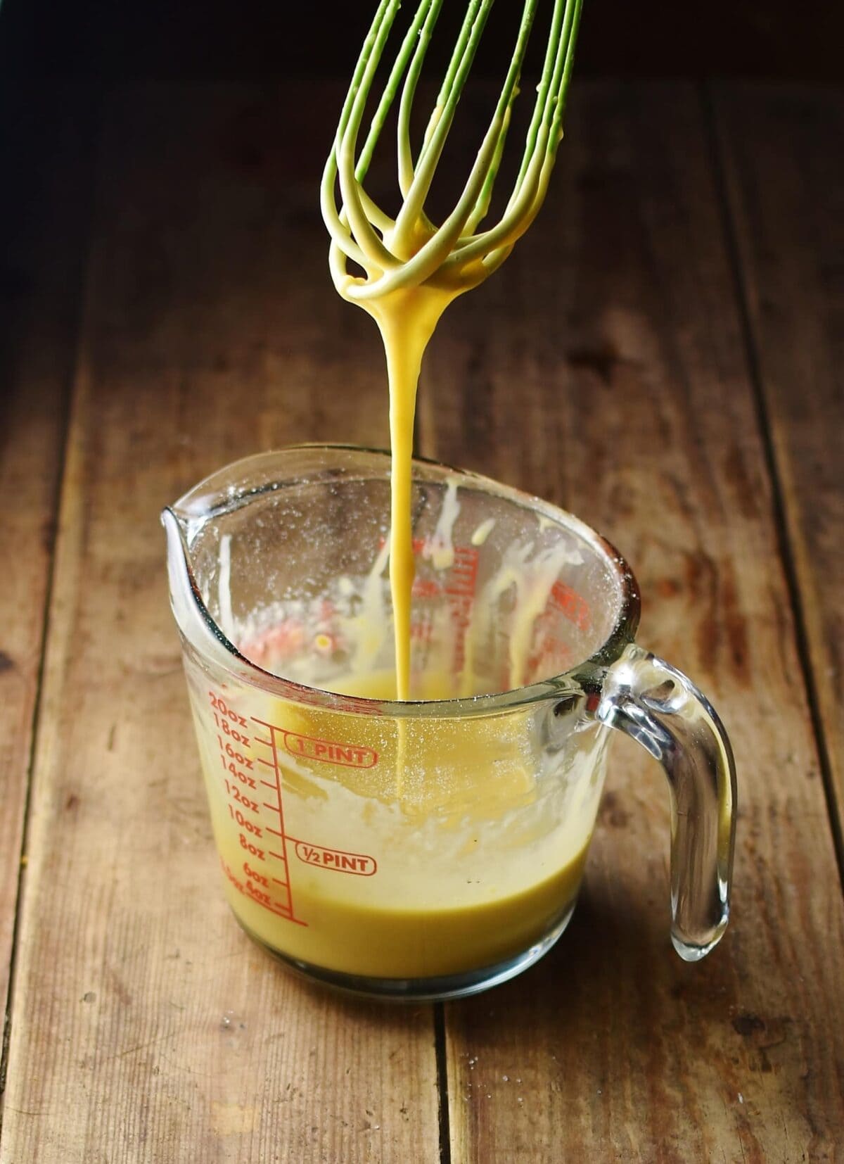 Egg mixture in measuring jug with mixture dripping from green whisk above.