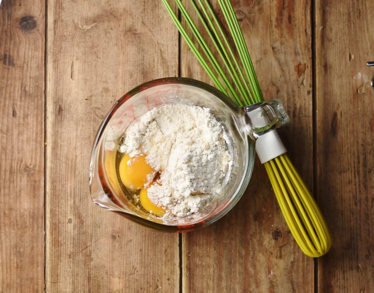 2 eggs and flour in jug with spout and green whisk to the right.