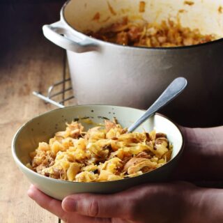 Sauerkraut stew in green bowl with spoon with hands holding it, and grey pot with stew in background.