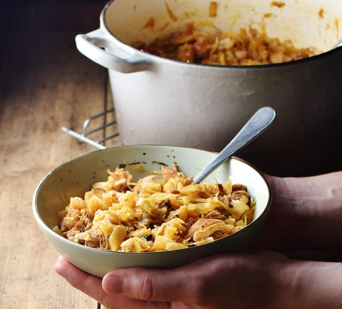 Sauerkraut stew in green bowl with spoon with hands holding it, and grey pot with stew in background.