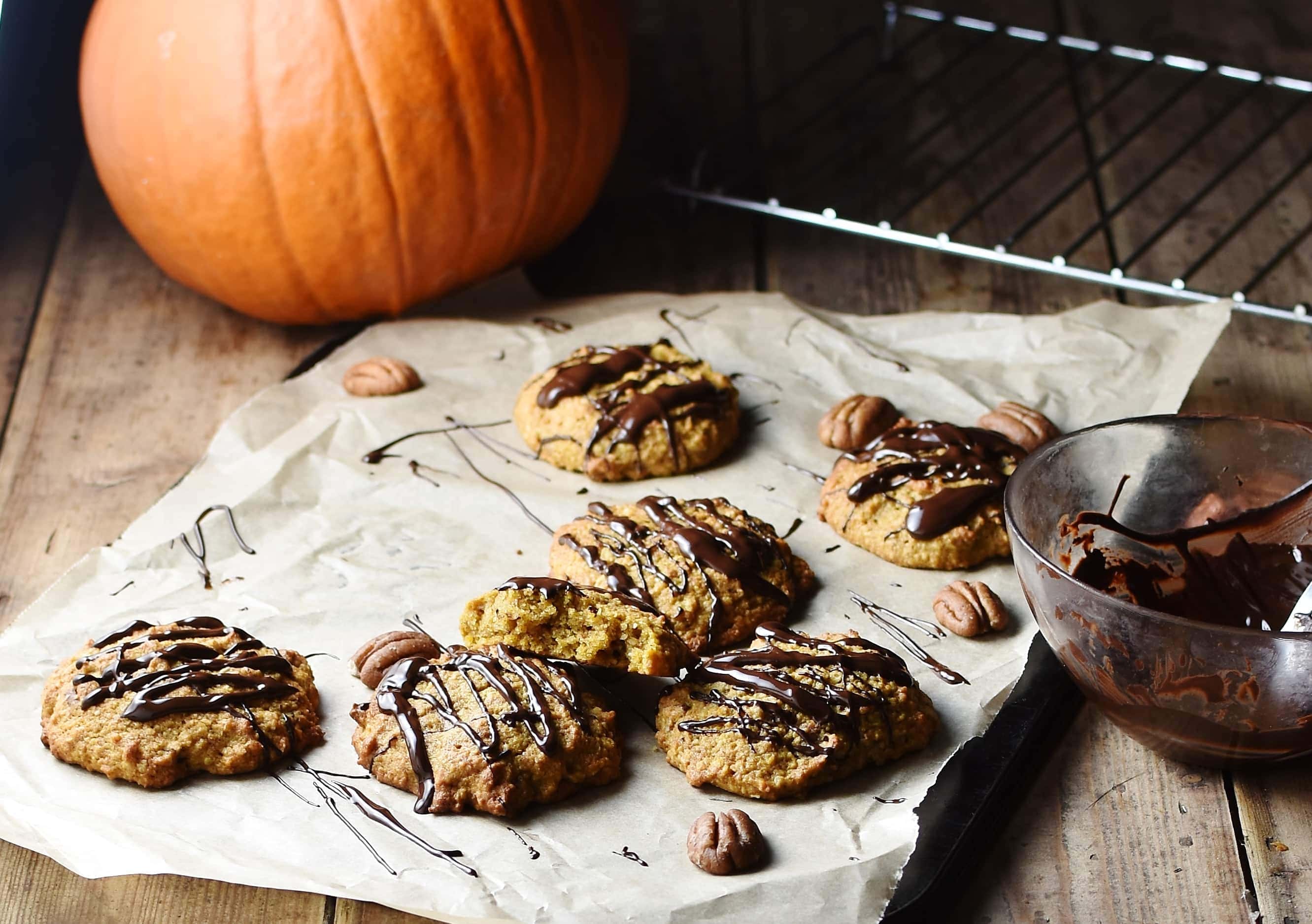Cookies with chocolate drizzle on paper with pecan nuts, and pumpkin in background.