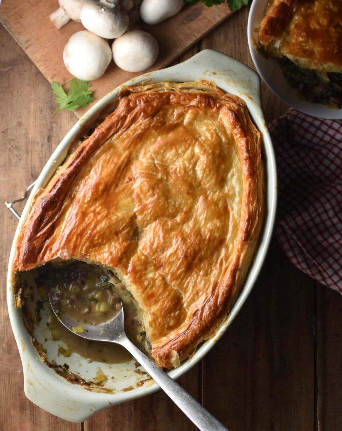Mushroom and leek pie topped with golden brown pastry in white oval dish with spoon, mushrooms and pie in white dish in background.
