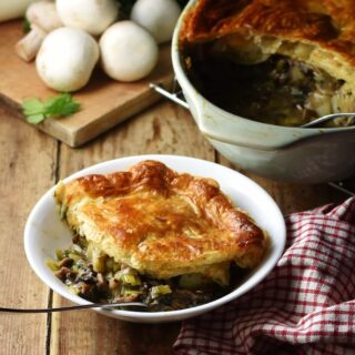 Side view of leek and mushroom pie topped with golden brown pastry on white bowl with fork, plaid red-and-white cloth to the right, pie in dish and mushrooms in background.