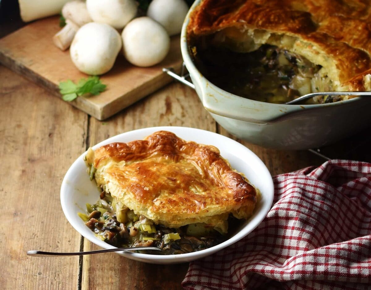 Side view of leek and mushroom pie topped with golden brown pastry on white bowl with fork, plaid red-and-white cloth to the right, pie in dish and mushrooms in background.