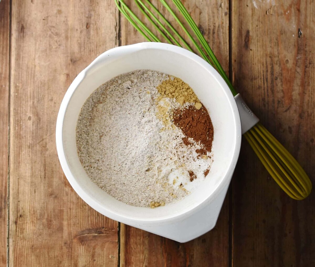 Flour and spice mixture in large white bowl with green whisk in background.