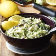 Side view of shredded brussels sprout slaw with lemon wedges and spoon in purple bowl on top of white plate, with lemon and zester in background.