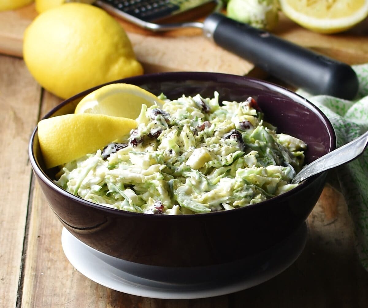 Side view of shredded brussels sprout slaw with lemon wedges and spoon in purple bowl on top of white plate, with lemon and zester in background.