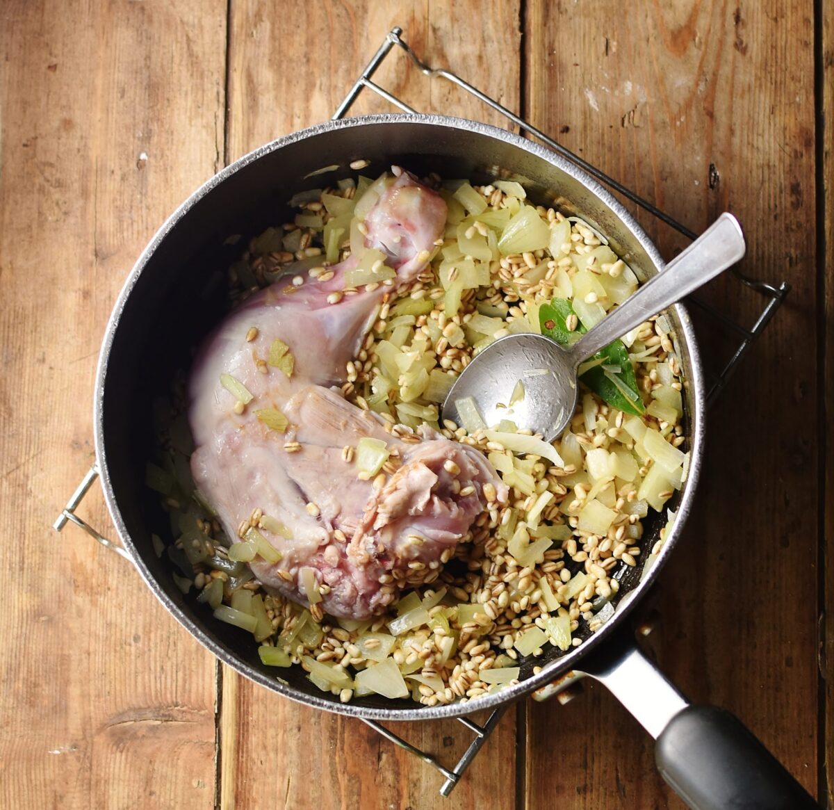 Top down view of chopped onions and barley with chicken leg and spoon in large pot.