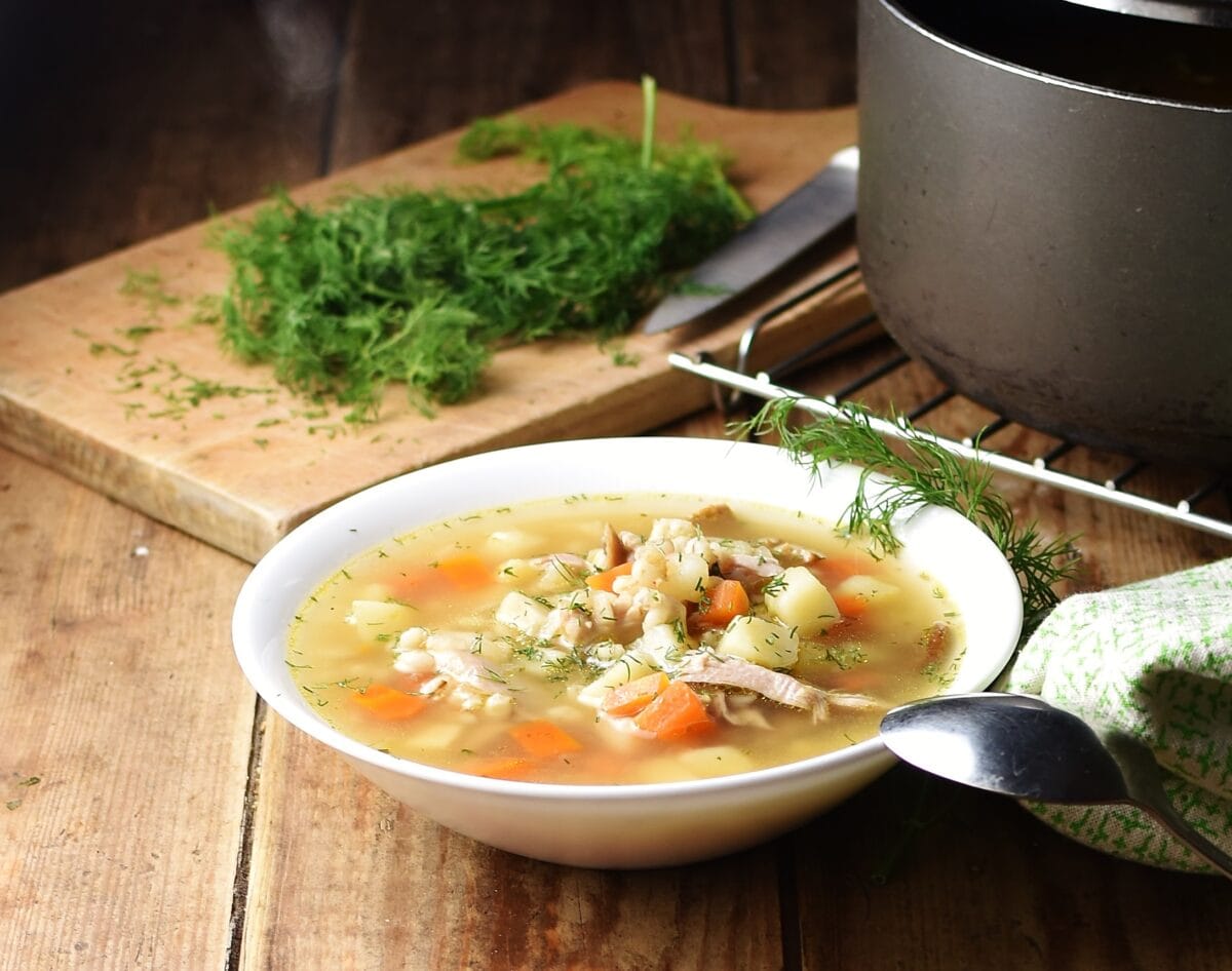 Side view of chunky barley soup in white bowl with spoon, fresh dill on wooden board and black pot in background.
