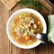 Chunky potato, carrot and barley soup in white bowl with spoon, green cloth to the right and dill at the top.
