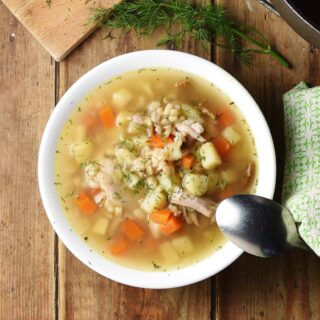 Chunky potato, carrot and barley soup in white bowl with spoon, green cloth to the right and dill at the top.