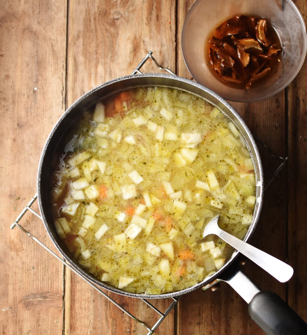 Chunky vegetable barley soup in large pot with spoon, and porcini mushrooms in small dish with water in top right corner.